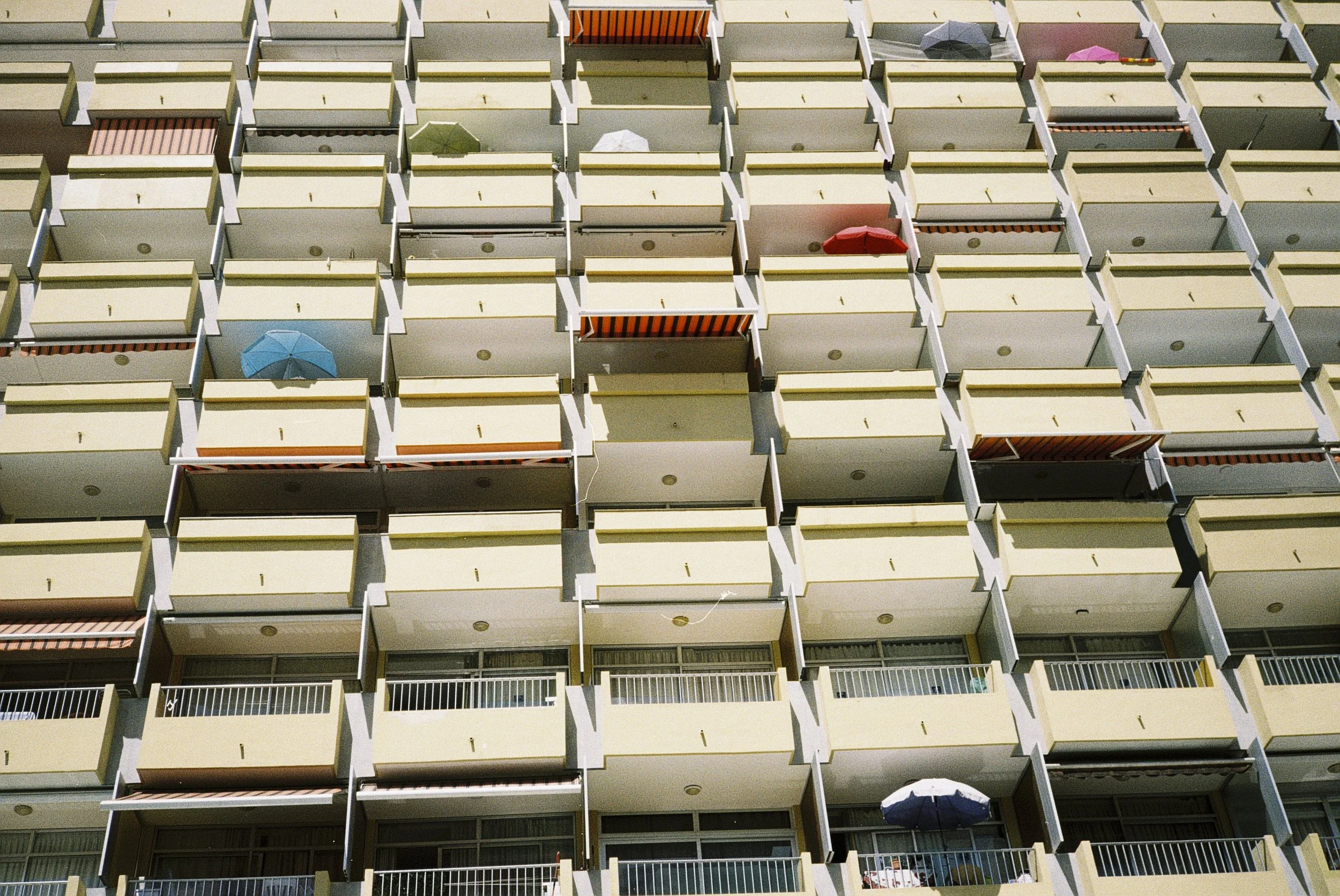 Mehrstöckiges Apartmentgebäude mit mehreren Balkonen, einige mit Sonnenschirmen unterschiedlicher Farben, darunter blau, grün, rot und pink.