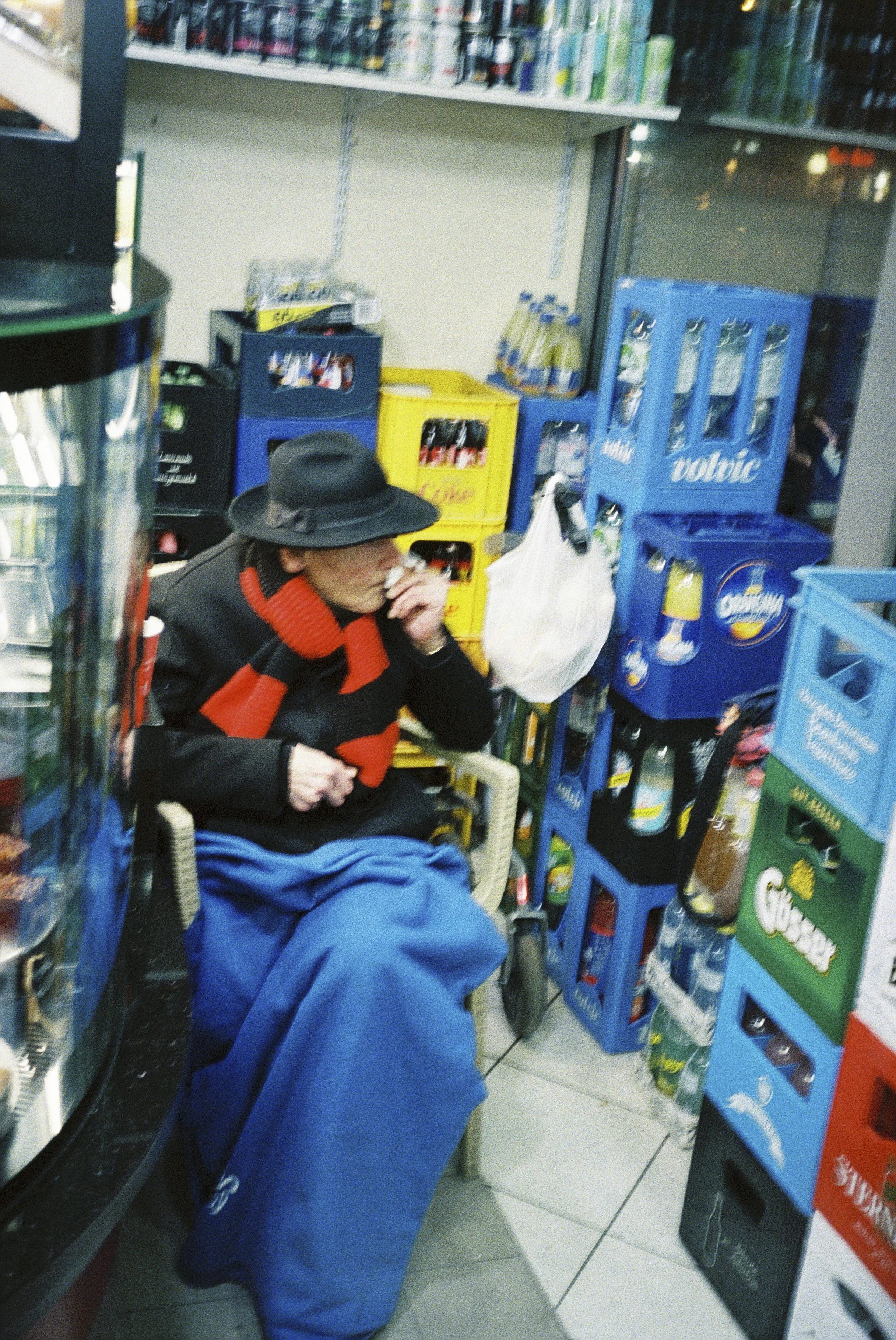 Eine ältere Frau sitzt in einem Supermarkt vor einem Regal mit Getränkekästen, trägt einen Hut und eine rote Strickjacke.