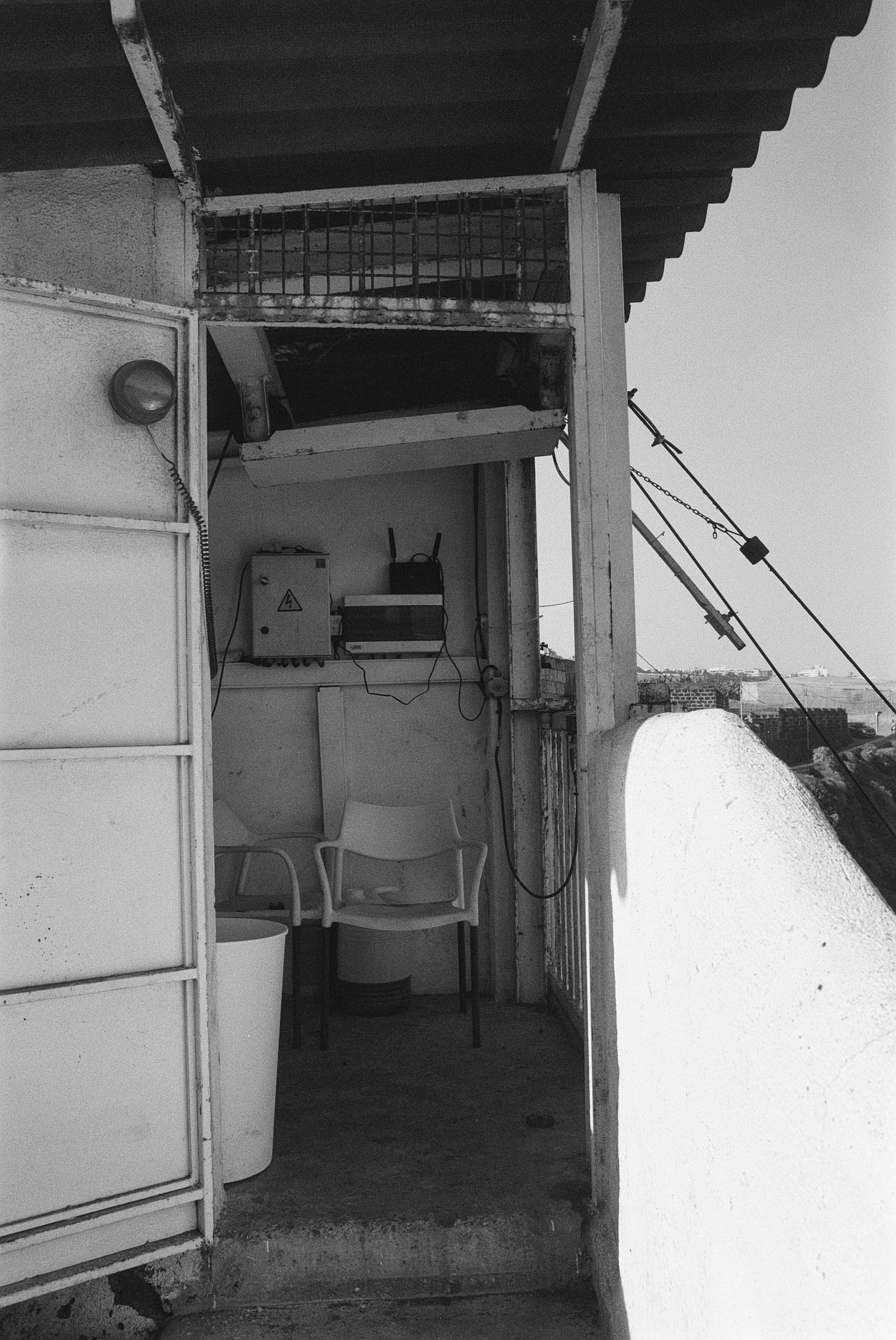 Blick in einen kleinen, abgeschlossenen Balkon mit Sitzgelegenheit, Wäscheleine und schattigem Dach, in schwarz-weiß Fotografie.