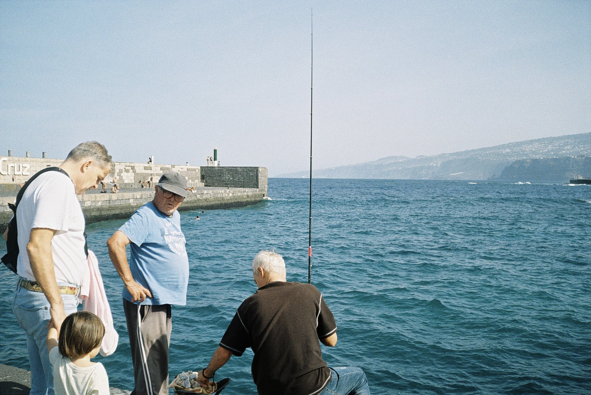 Gruppe von Menschen, die am Wasser stehen, mit Blick auf das Meer und den Himmel im Hintergrund. Einige sind im Wasser, während andere am Ufer stehen.
