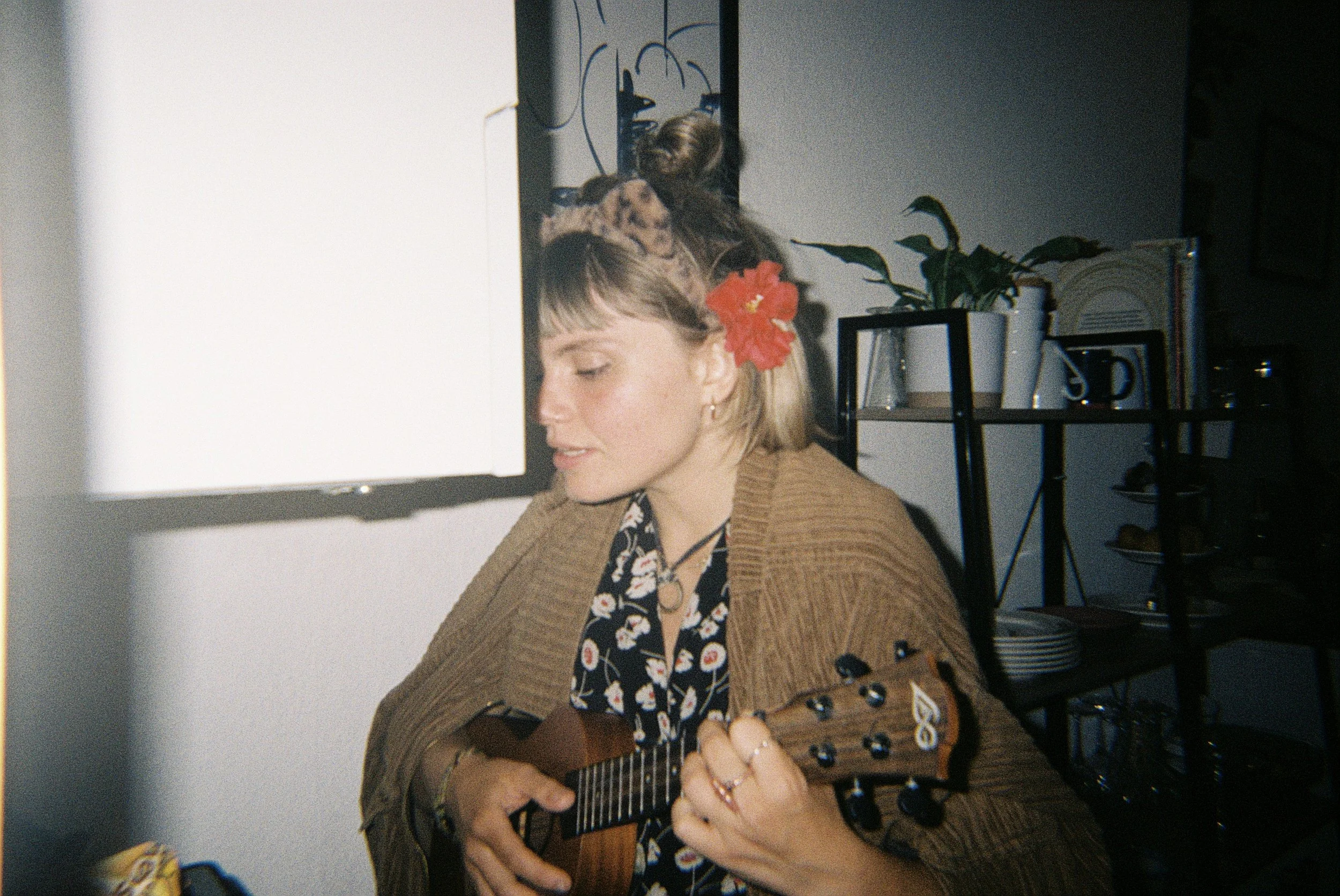 Junge Frau mit Petticoat-Kleid und roten Hibiskusblume im Haar spielt Gitarre in einem Raum mit Pflanzen und Büchern.