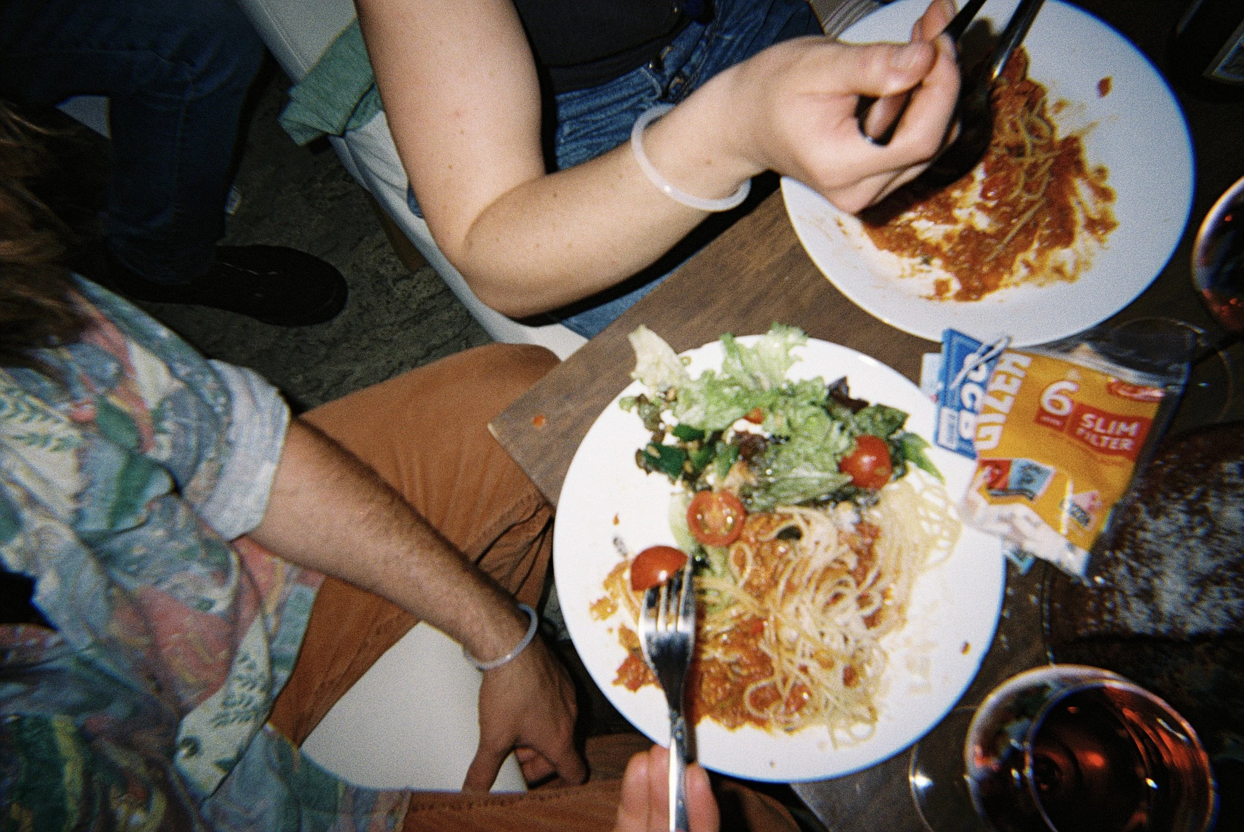 Person isst eine Portion Spaghetti mit Tomatensauce, daneben ein grüner Salat, am Tisch stehen Gläser mit rotem Wein.