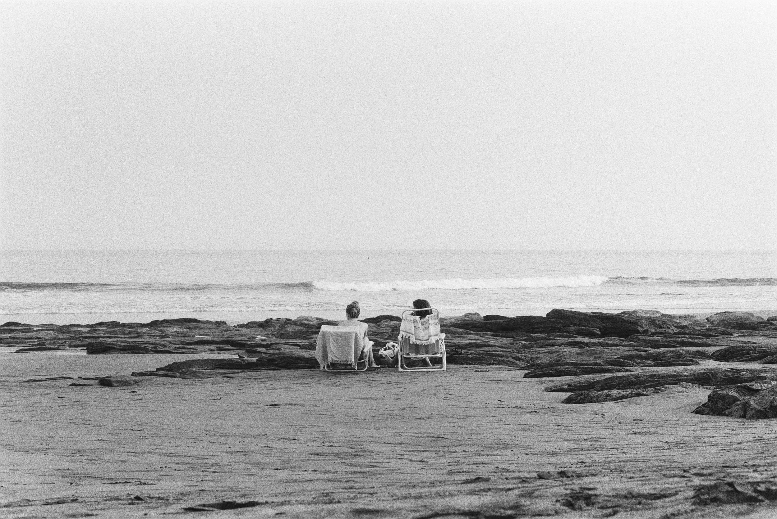 Zwei Menschen sitzen auf der Tag oder Nacht an einem Strand, umgeben von Felsen, im Meer. Es ist eine ruhige, monochrome Szene.