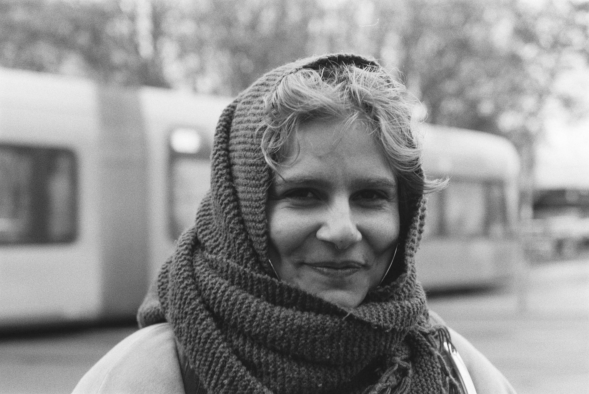 Nahaufnahme eines lachenden jungen Mannes mit blondem, lockigem Haar, der einen karierten Hut trägt und eine dicke Strickschlauch um den Hals hat, im Freien mit verschwommenem Hintergrund.