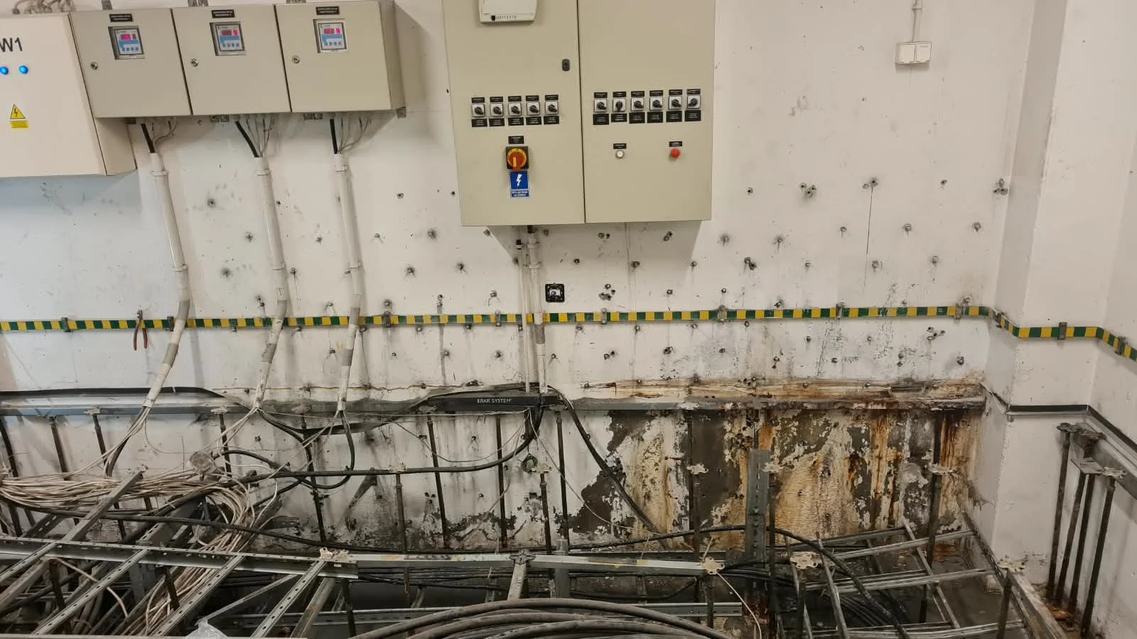 Interior of an unfinished or decommissioned electrical or data cabling room, with many cables and wiring on the floor, electrical boxes and panels on the wall, and visible damage to the wall and floor.