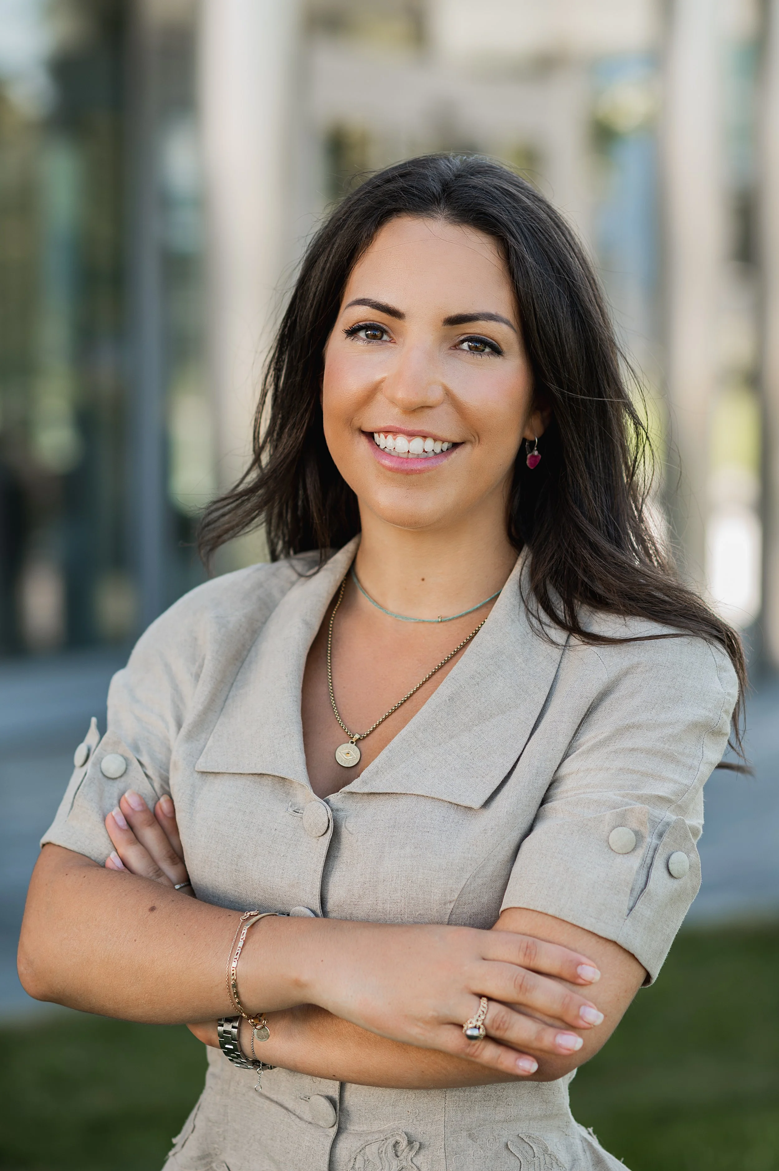 A woman with dark hair and a light beige blazer smiling outdoors with her arms crossed.