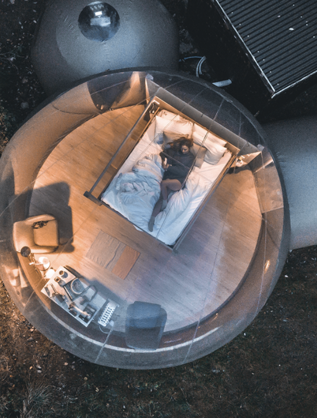 A person lying inside a transparent, bubble-shaped bedroom with wooden floors. The room contains a bed, a small table with electronic equipment, a chair, and a lamp, surrounded by an outdoor environment.