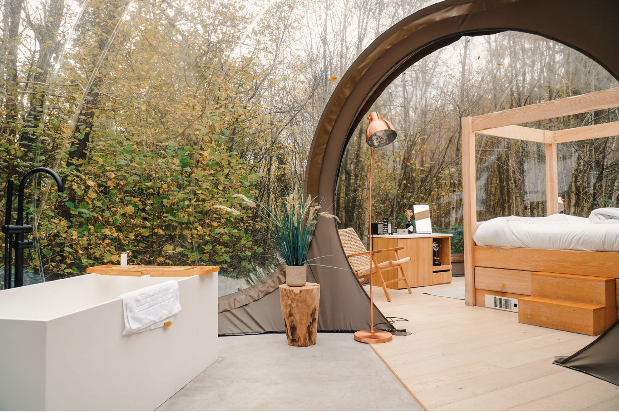 Interior of a cozy glamping tent with a wooden bed, a small table, a reading lamp, and a bathtub, with large windows showing autumn trees outside.