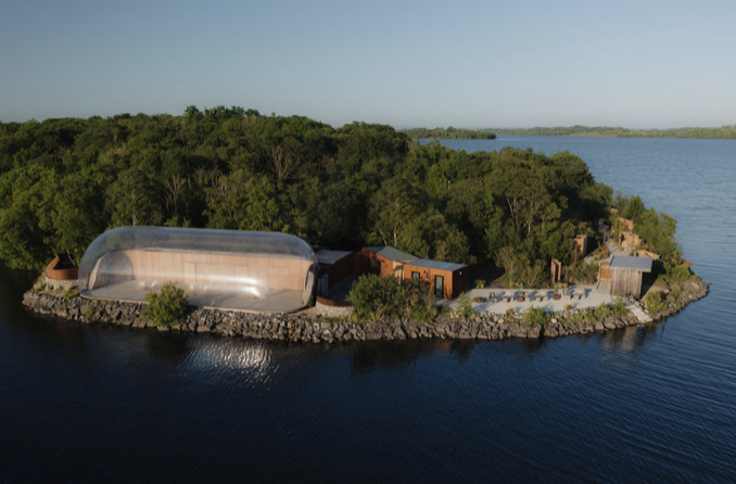 An island with a large transparent dome structure, surrounded by water and lush green trees.