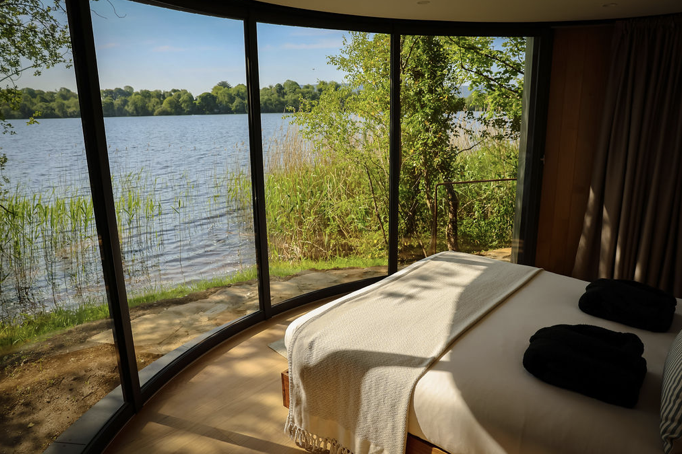 A bedroom with a curved glass wall and a view of a lake with trees and reeds outside, a bed with white bedding and black pillows, and sunlight casting shadows inside.