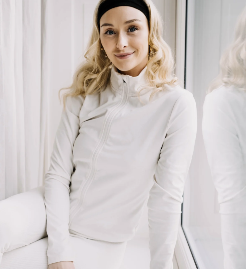 A woman with blonde wavy hair wearing a black headband and white athletic jacket, standing next to a mirror, smiling gently.