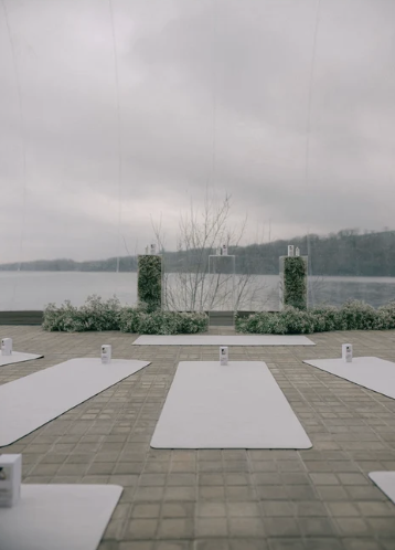 Outdoor yoga setup on a dock by a lake with white mats and small lanterns, cloudy sky and distant shore in the background.