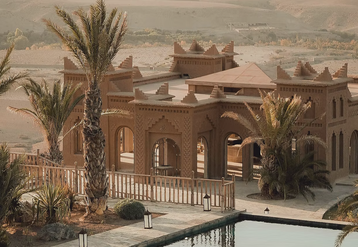 A desert-style house with traditional Moroccan architecture, surrounded by palm trees and overlooking a landscape. There is a pool in the foreground and lanterns along the pathway.