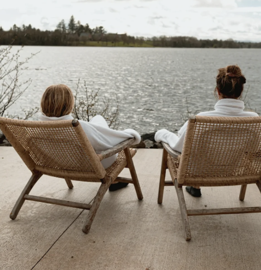 Two people seated in wicker chairs outdoors by a lake with trees in the background, relaxing and enjoying the view.