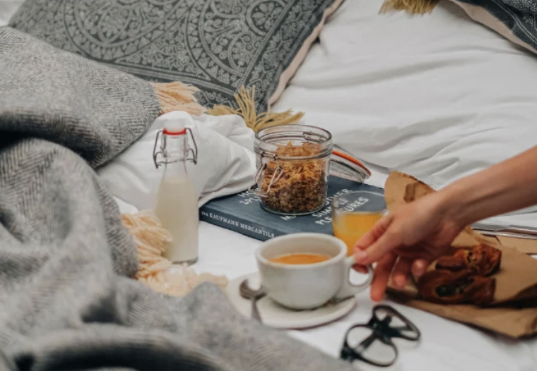 A cozy bed with breakfast items including a cup of coffee, a glass of orange juice, a bottle of milk, a jar of cereal, and some bread. There are also a pair of glasses and a book on the bed.