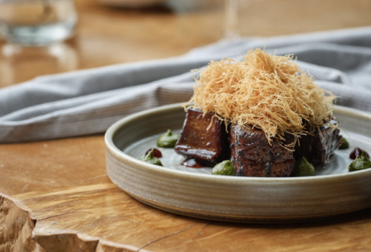 A plated dish with braised meat topped with crispy shredded garnish, served with green beans and sauce on a ceramic plate.