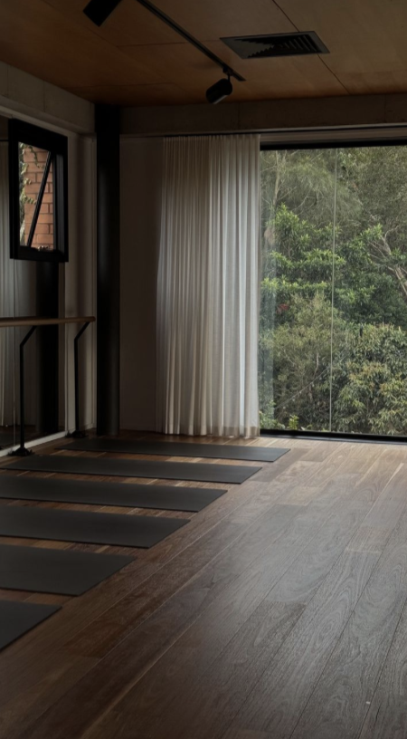 An interior of a yoga or meditation room with wooden flooring, black yoga mats, a window showing greenery outside, and white curtains.