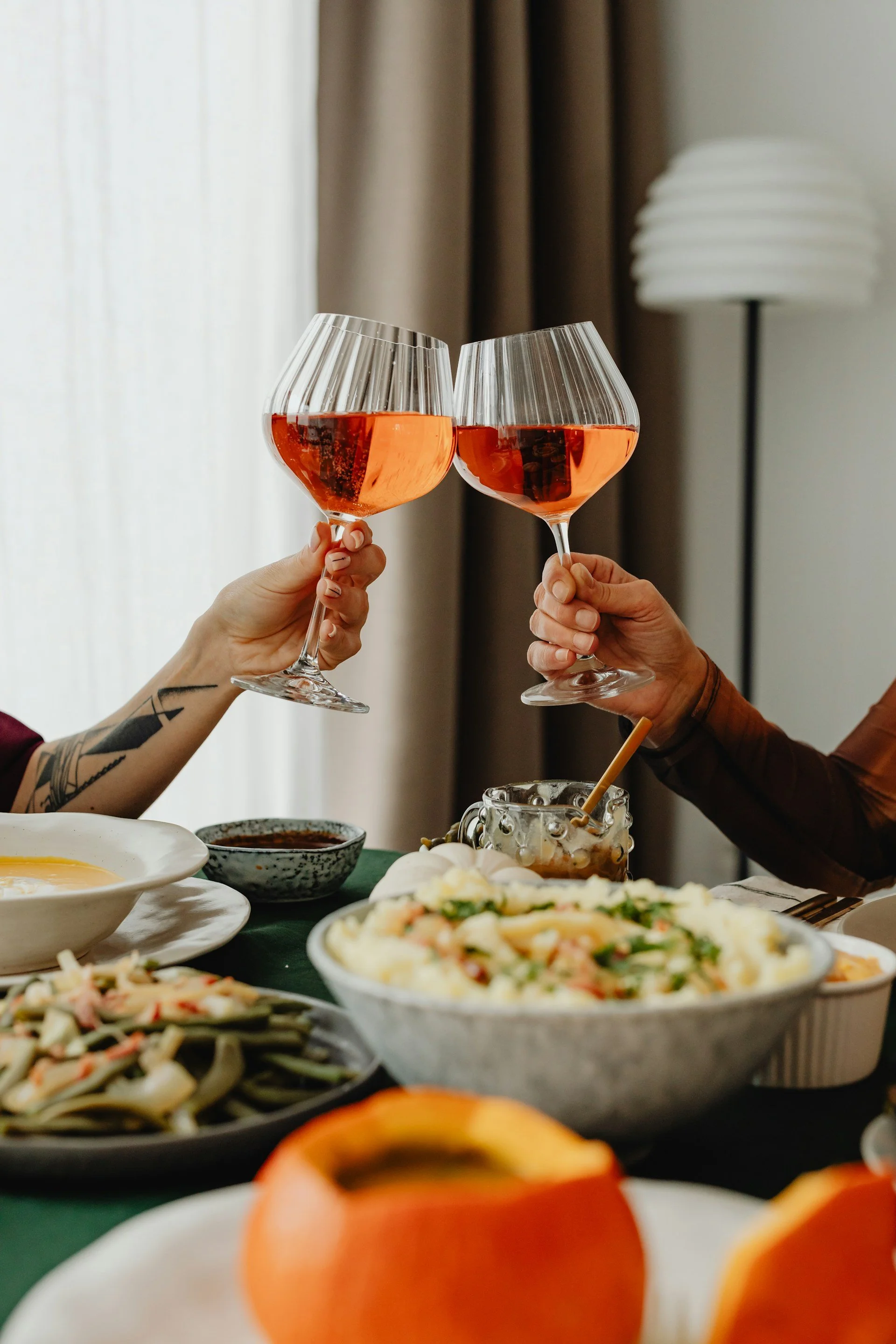 Two people holding glasses of rosé wine clinking together over a table with various dishes including salads, pasta, and appetizers in a cozy, well-lit dining setting.