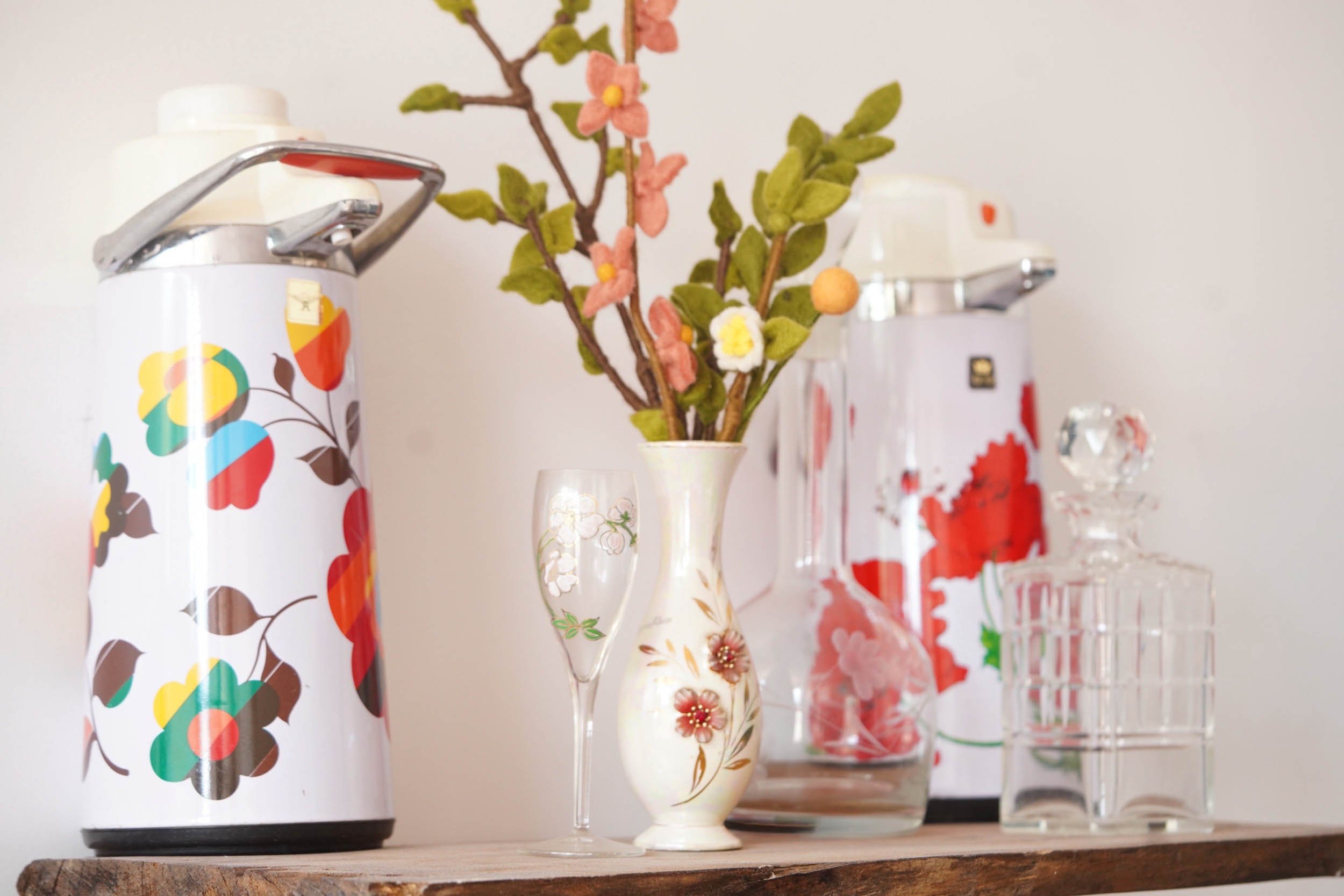 Collection of decorative glass vases and containers on a wooden shelf, some with floral or fruit patterns, displaying artificial flowers and small decorative items.