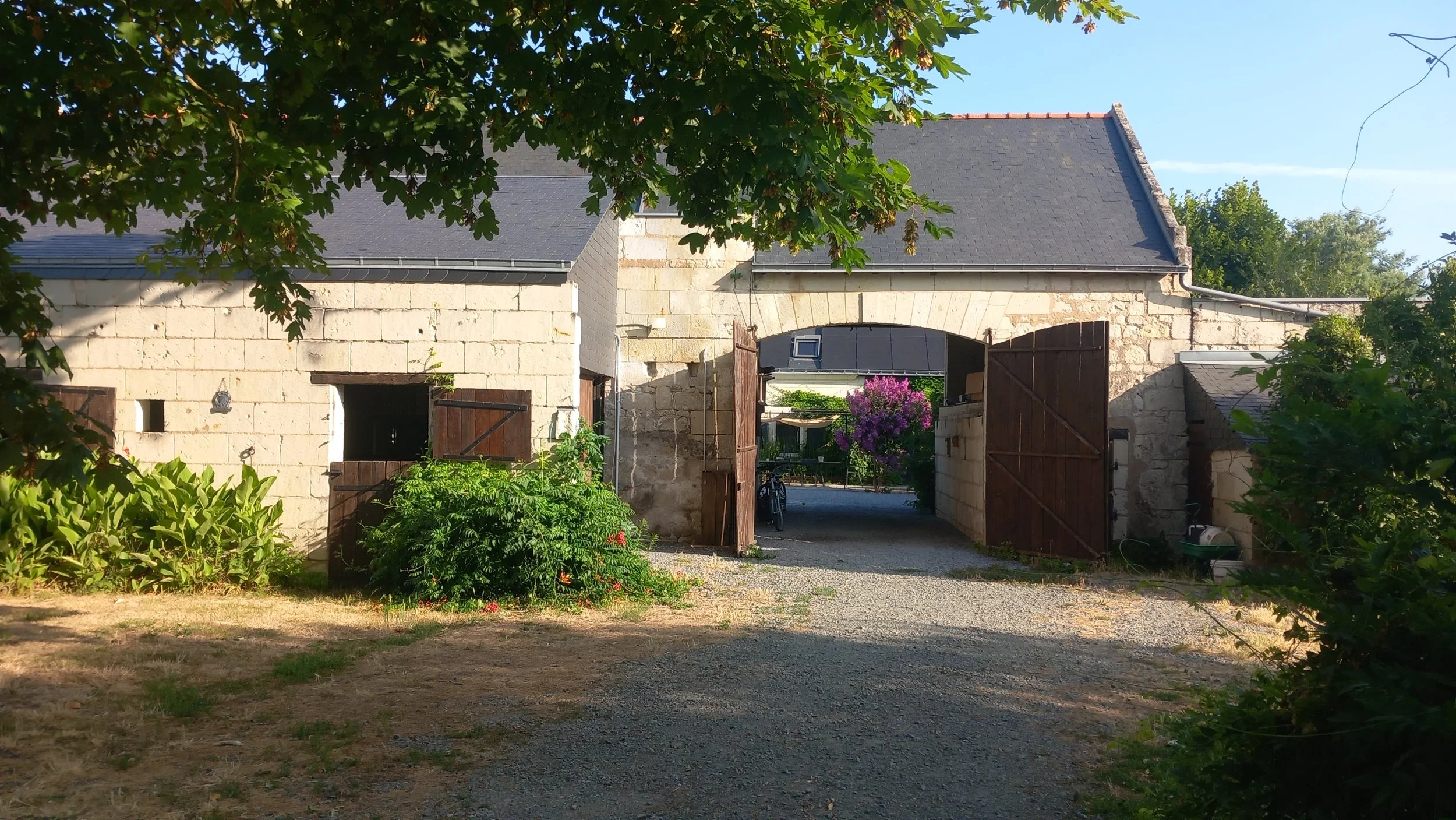 Une vieille ferme en pierre avec un portail en bois, entourée de jardin et d'arbres, sous un ciel bleu clair.