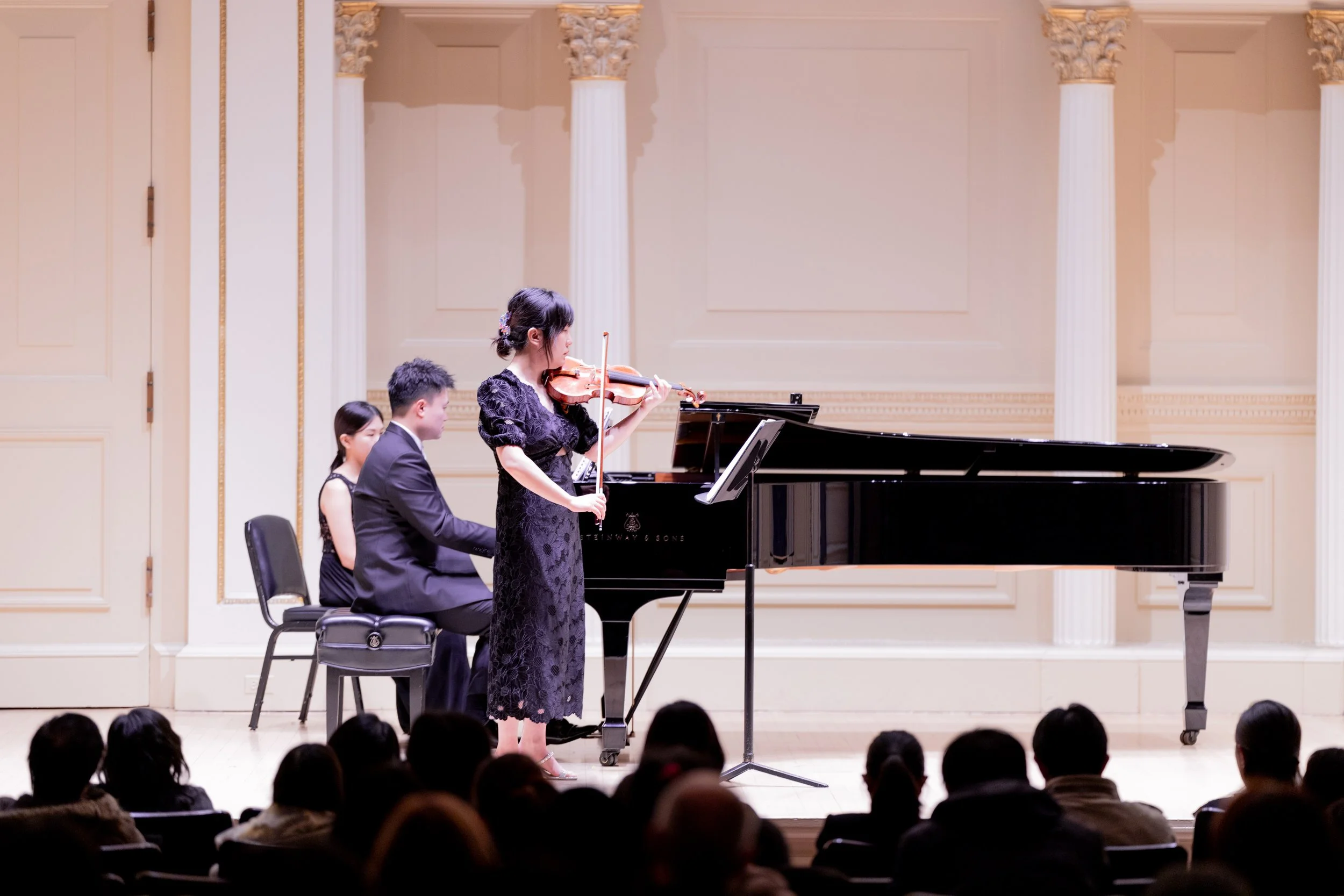 A classical music performance in a concert hall with four musicians: a woman playing the violin, another woman in the background, and two men playing the piano.
