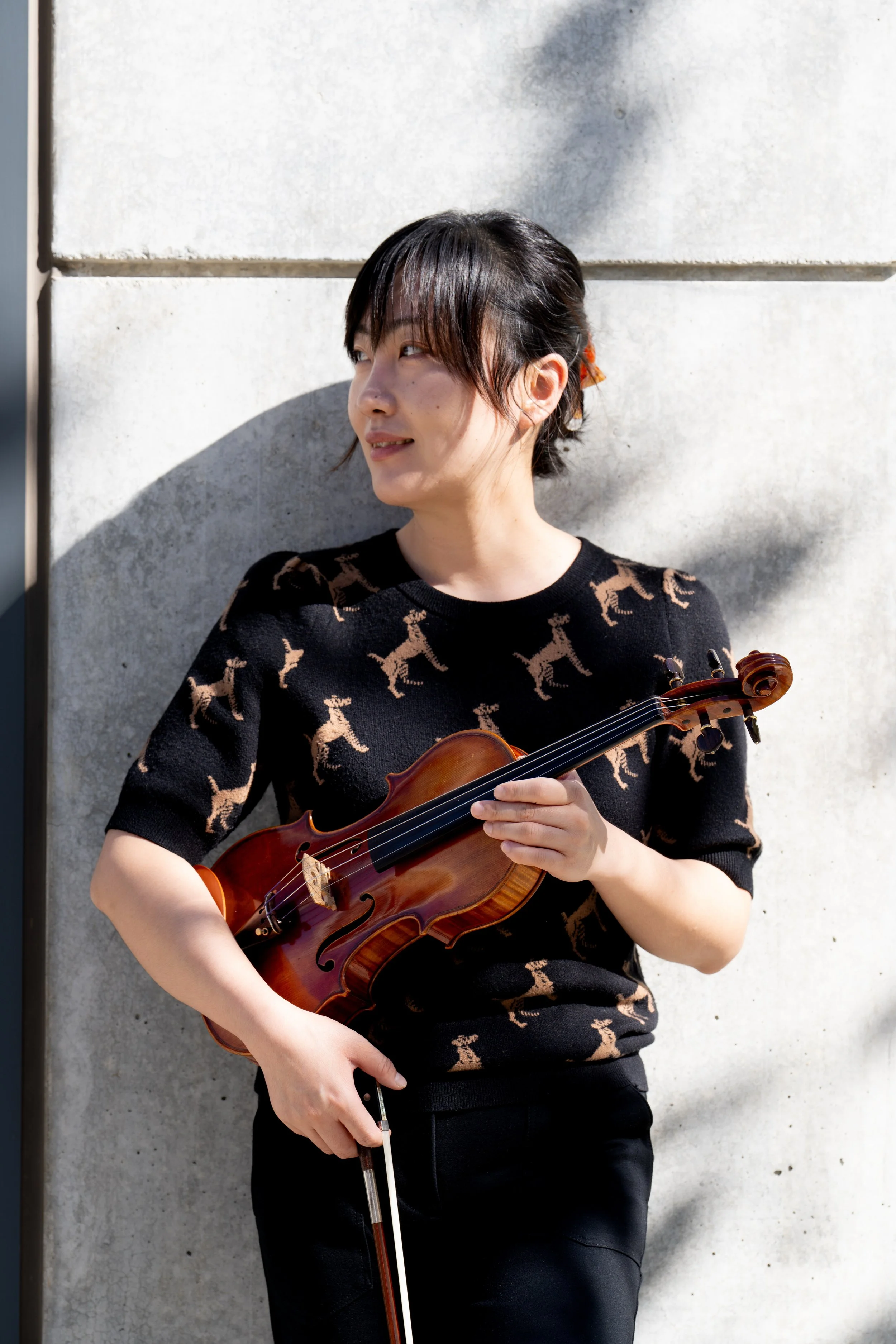 A woman with dark hair and light skin color is standing outdoors, holding a violin and a bow, with a light-colored concrete wall behind her. She is wearing a black short-sleeved shirt with a pattern of tan dogs on it.