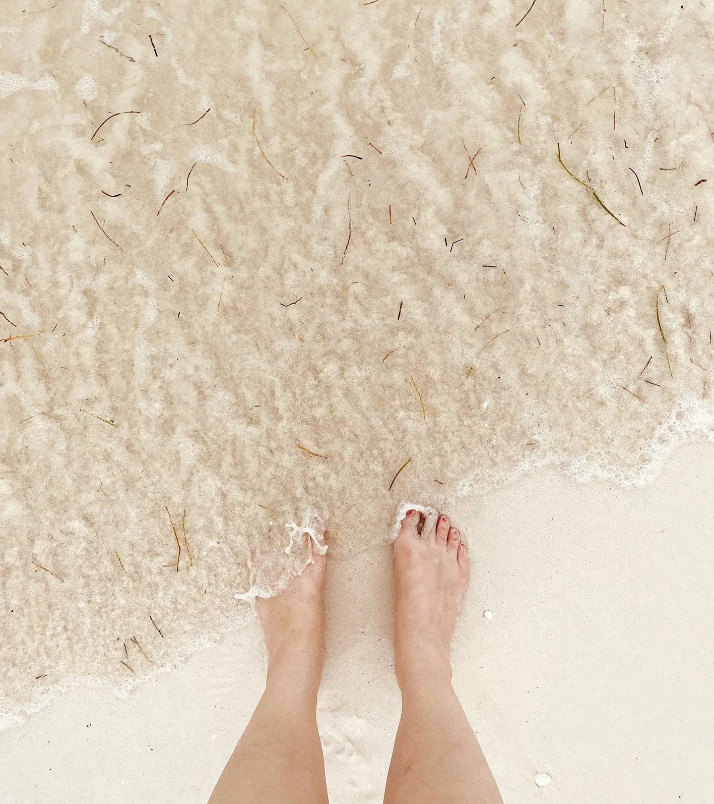 Person's feet standing in the shallow surf at the beach, with frothy ocean water and small seaweed fragments around.