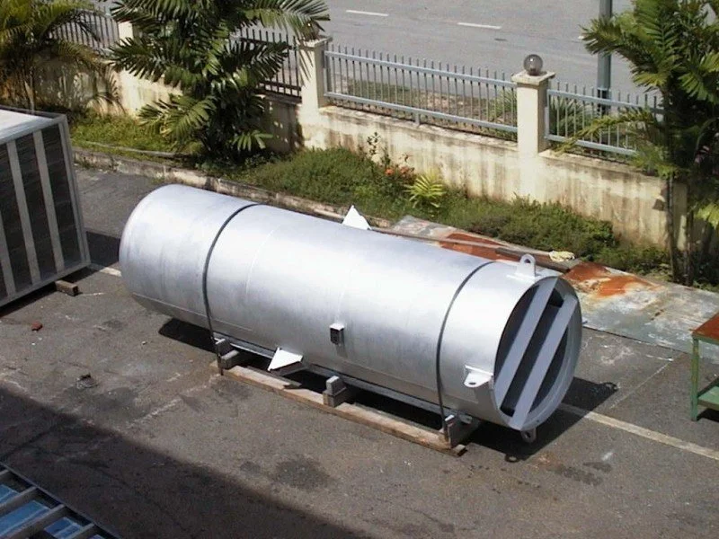 A large silver cylindrical tank lying on its side on a pallet on a rooftop, with surrounding plants and a metal fence in the background.