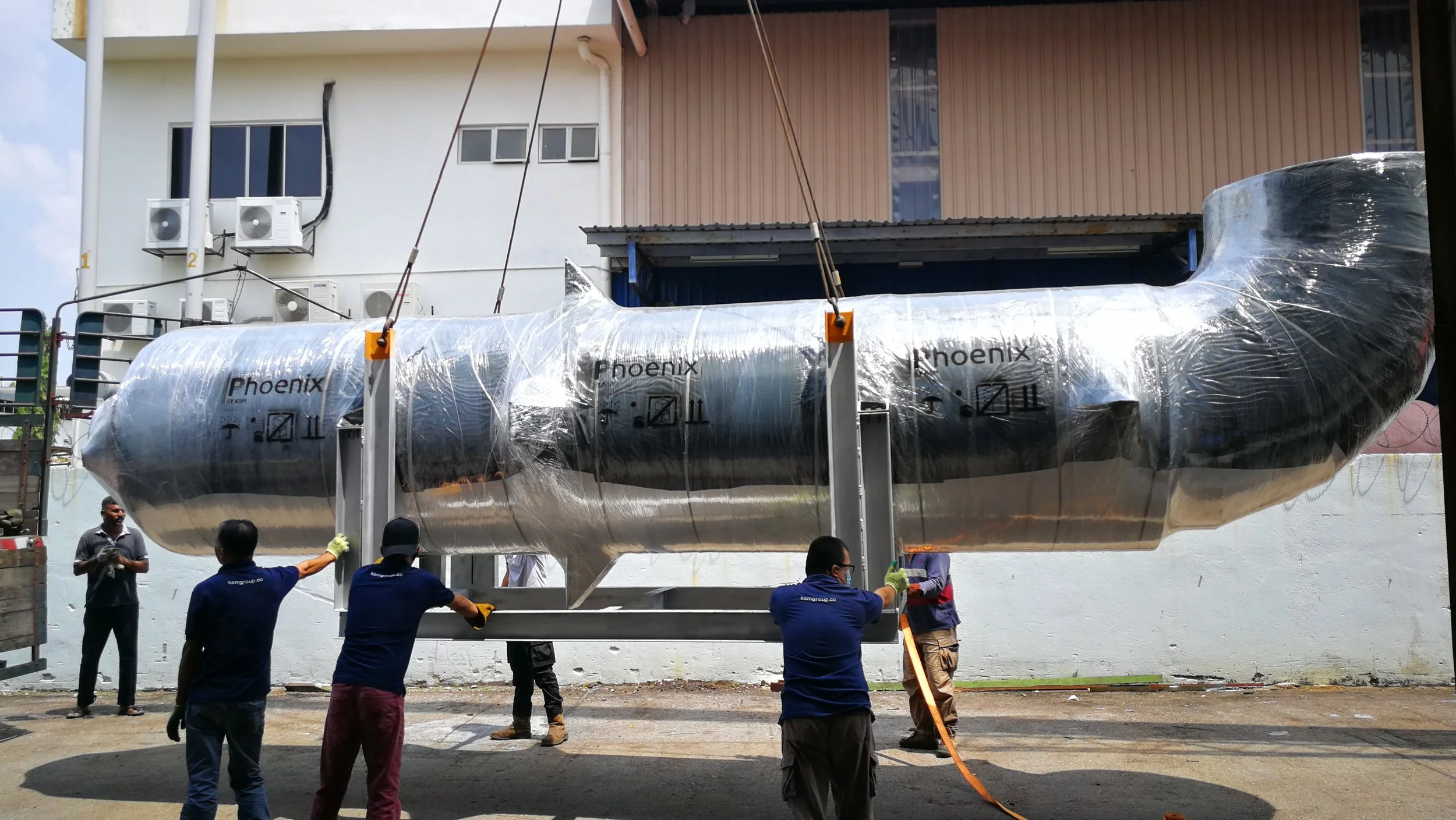 A large, shiny, silver submarine with the word 'Phoenix' written on its side is being lowered or lifted by several workers using straps and a crane outside a building.
