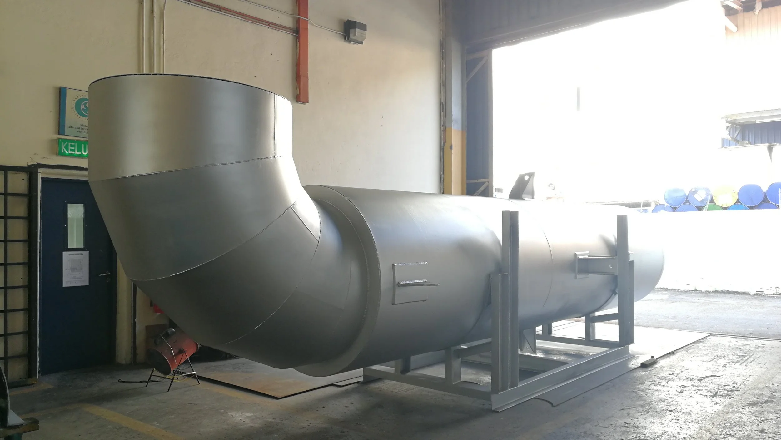 A large industrial metal vessel or tank inside a warehouse or factory, with a curved top and cylindrical body, positioned horizontally on a support frame near an open garage door allowing natural light to enter.