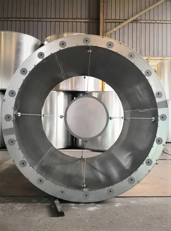 Large industrial metal coil with a hollow center, mounted in a factory setting with metal tanks in the background.