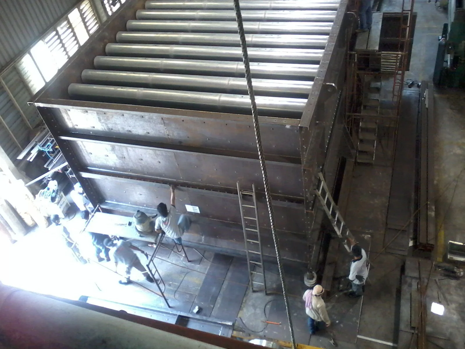 Workers assembling a large metal structure with roller tubes and scaffolding inside an industrial warehouse.