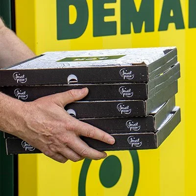 Person holding four pizza boxes stacked in front of The Spice Co. pizza machine.