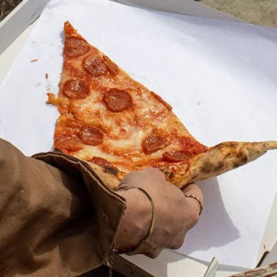 A person's hand holding a large slice of pepperoni pizza on white paper.