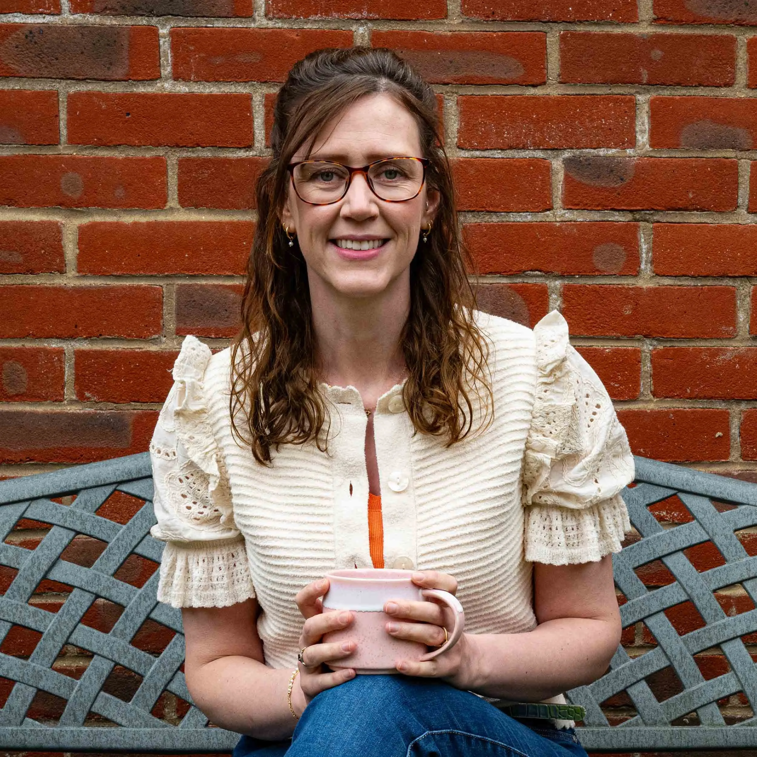 Woman with glasses holding a cup, sitting on a bench in front of a red brick wall.