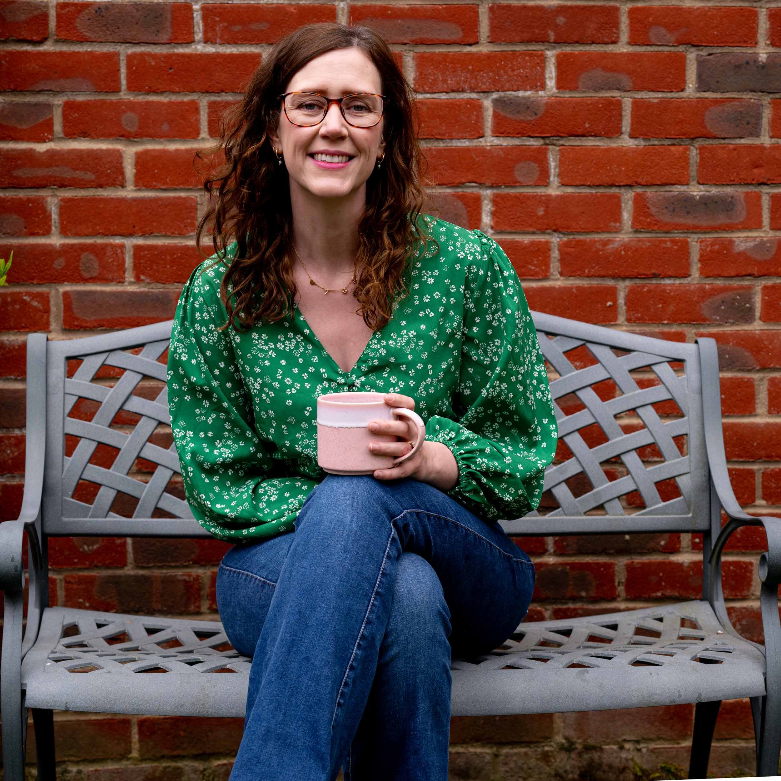 A woman with long wavy brown hair, wearing glasses, a green floral blouse, and blue jeans, sits on a gray metal bench holding a pink mug with both hands, in front of a red brick wall.