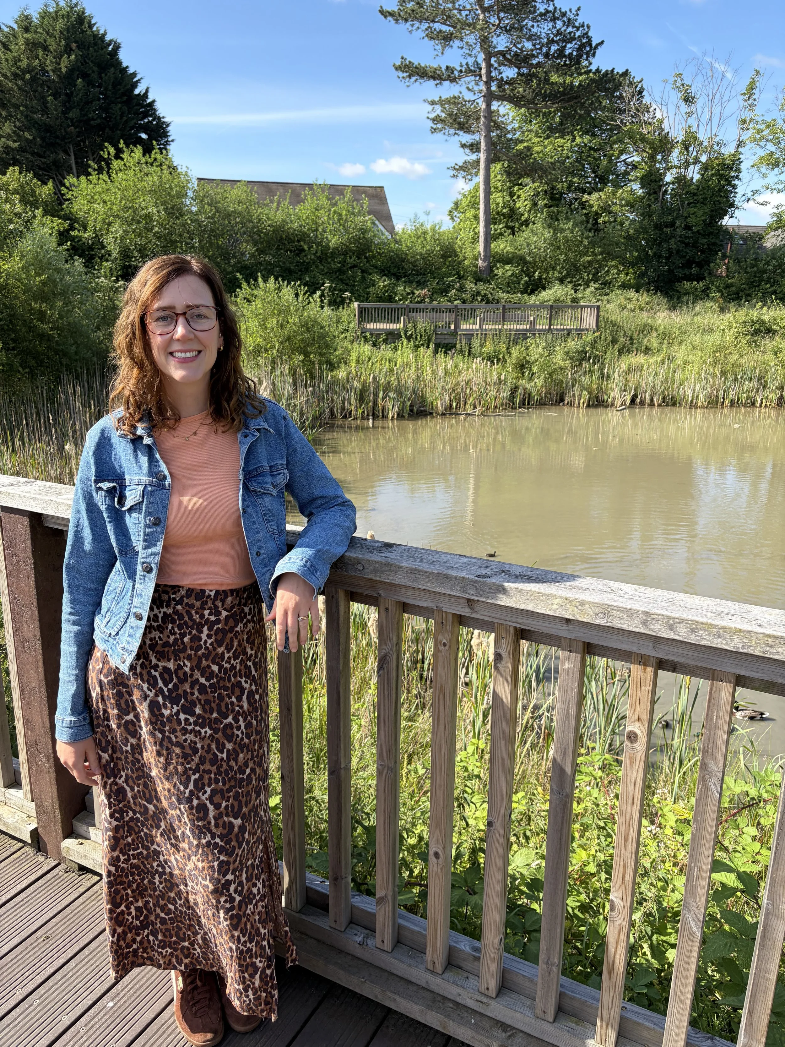 A woman with glasses standing on a wooden walkway next to a pond with greenery and trees in the background on a sunny day.