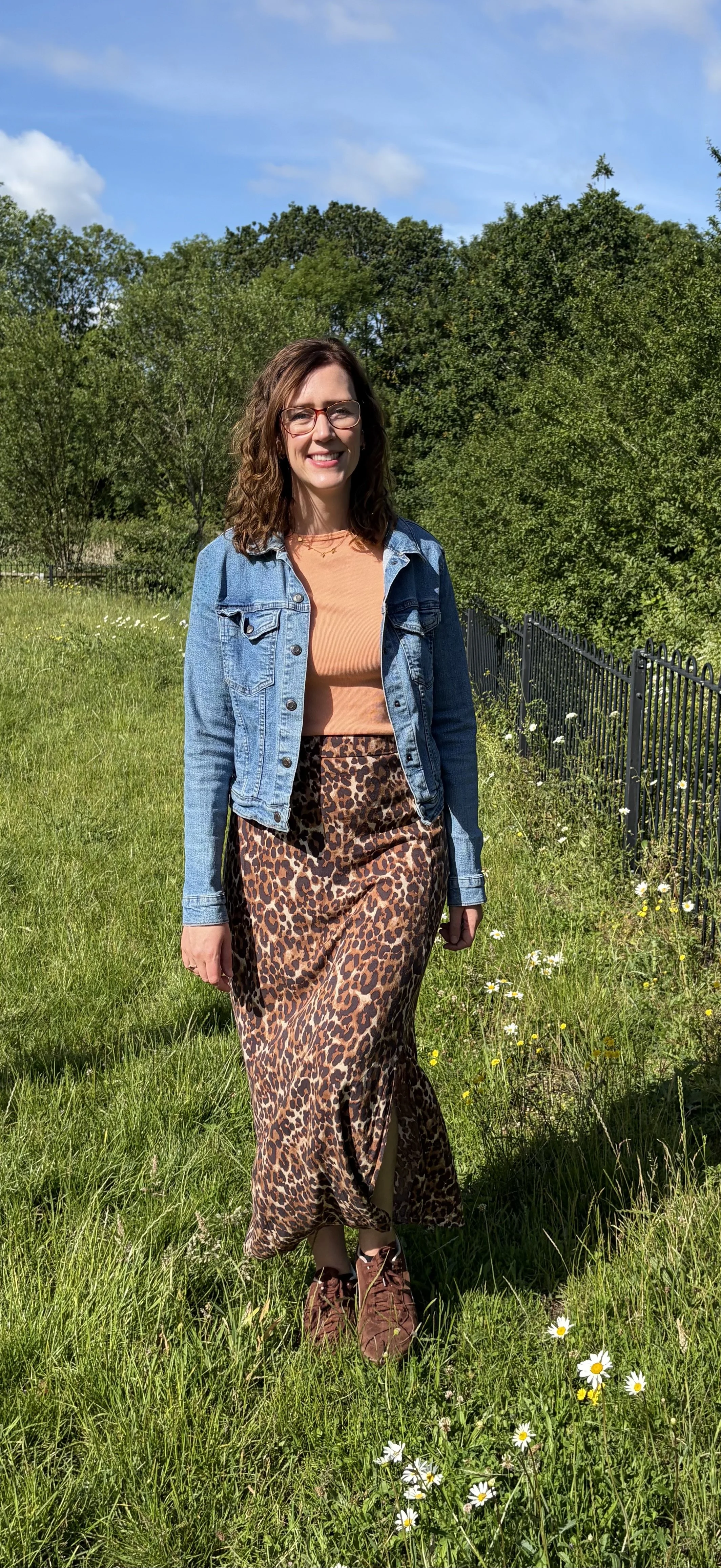 A woman standing outdoors on a sunny day, wearing a denim jacket, peach top, and leopard print skirt, with green trees and a blue sky in the background.