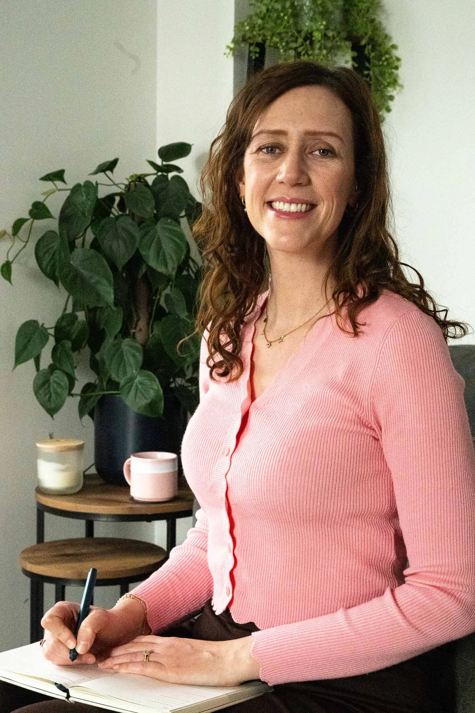 A woman with wavy brown hair, wearing a pink cardigan with red piping, is seated at a desk writing in a notebook with a black pen. Behind her, there is a potted plant on a black stand and a side table with candles and a pink mug. She is smiling and looking directly at the camera.
