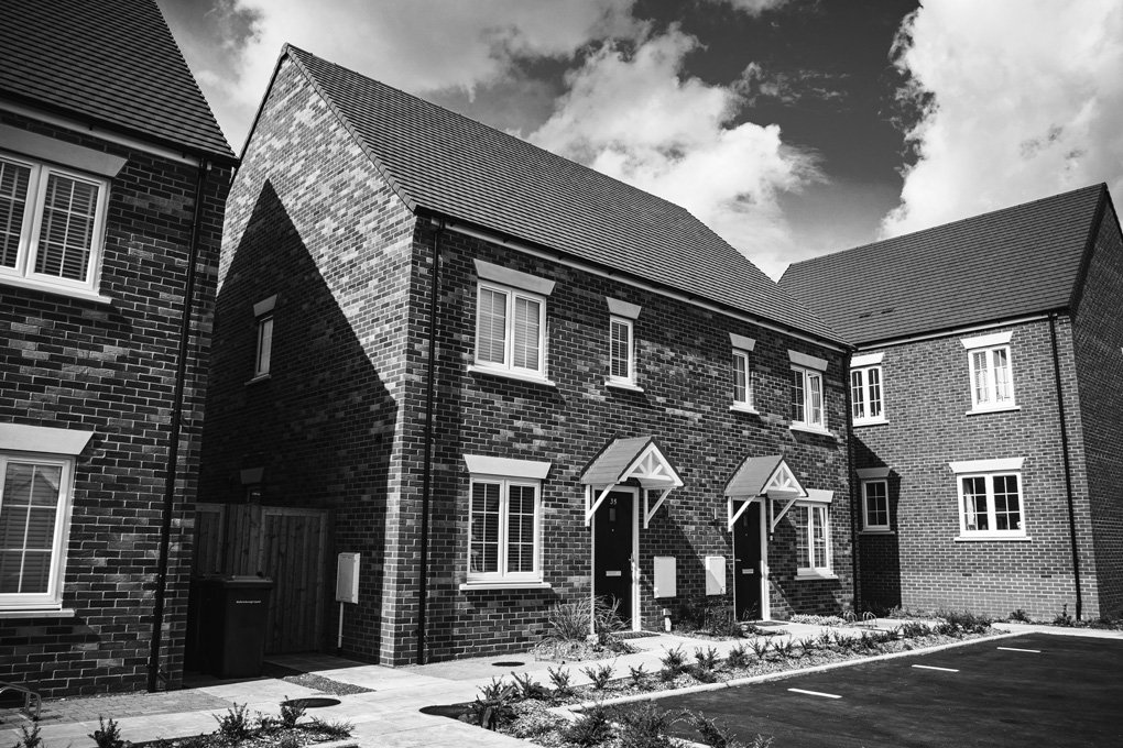 Black and white photo of modern brick townhouses with pitched roofs, small front gardens, and parking spaces in front. Some windows have white trim, and the sky above is partly cloudy.