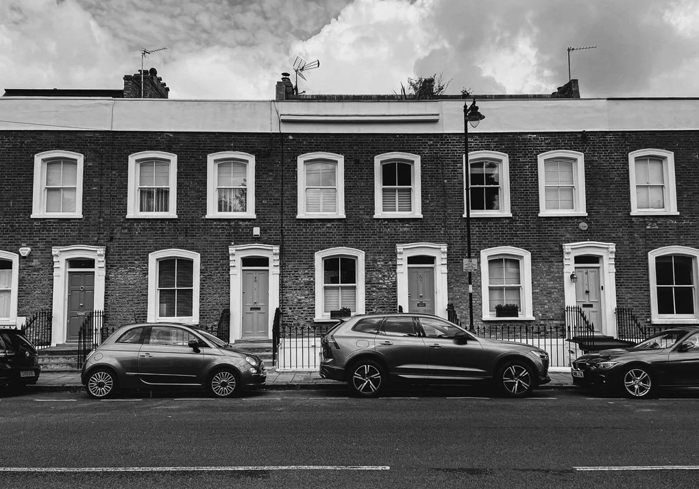 Row of three-story brick townhouses with front doors and windows, parked cars along the street, cloudy sky, black and white photograph.