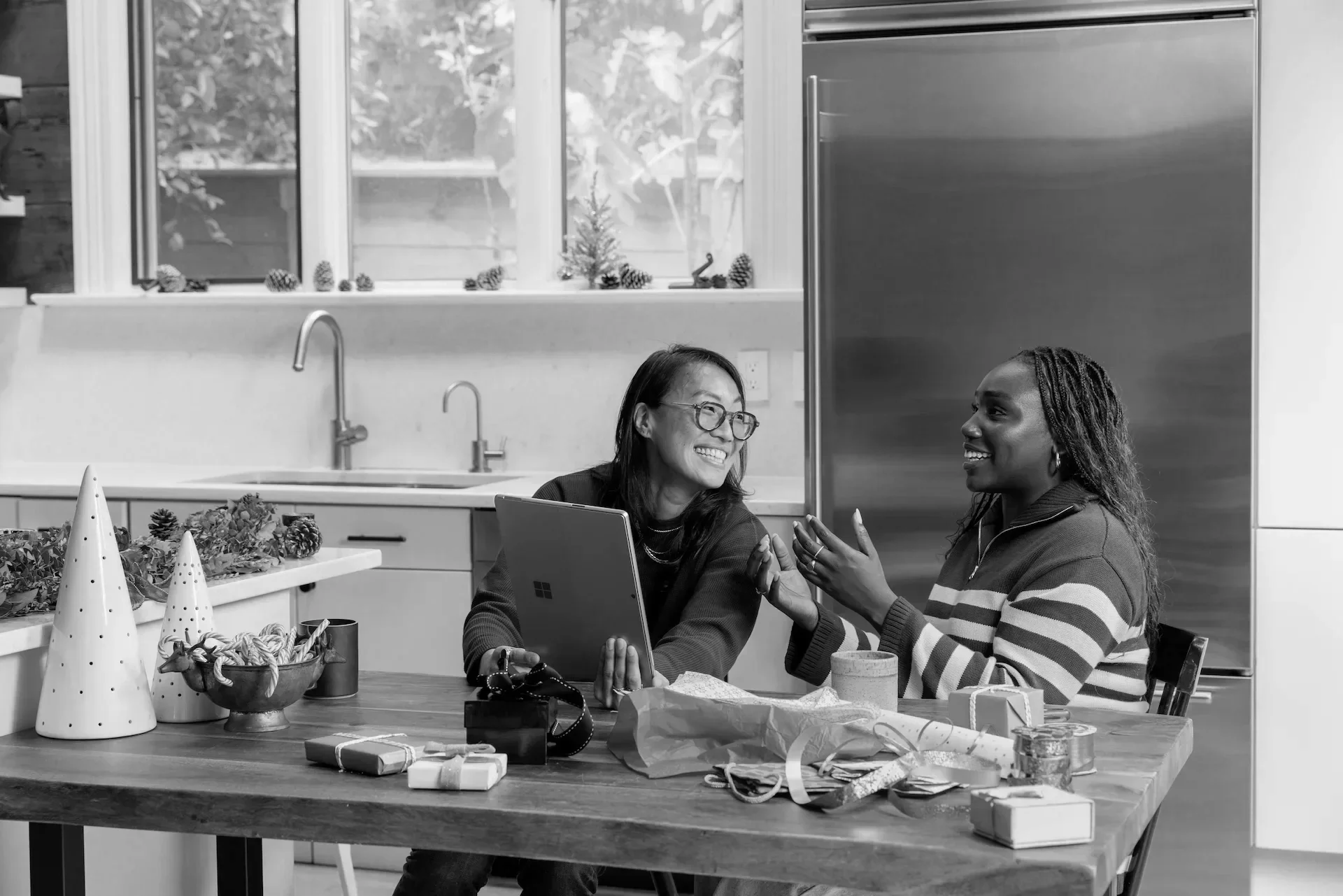 Two women are sitting at a table in a kitchen, smiling and talking. The table has Christmas decorations, wrapped gifts, and craft supplies. One woman is holding a tablet, and they appear to be engaged in a friendly conversation.