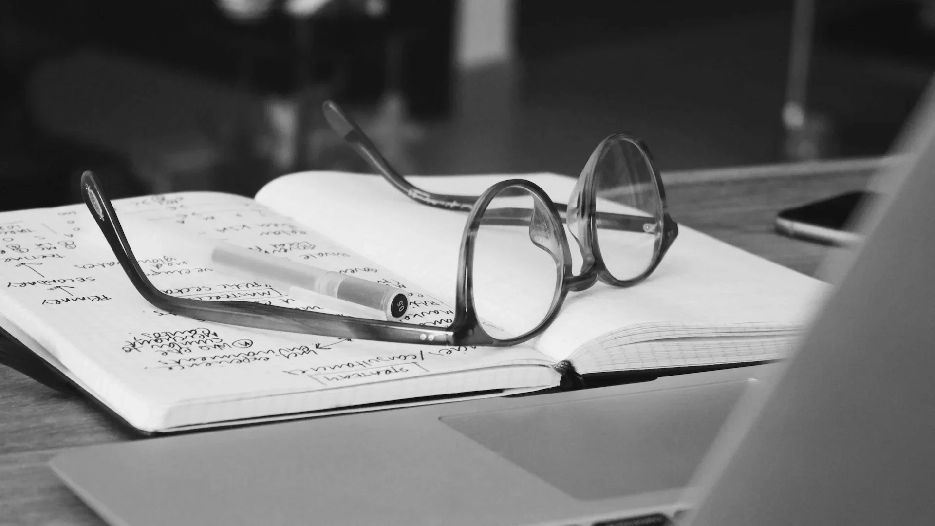 Black and white photograph of glasses, an open notebook with handwritten notes, a pen, and a partial view of a laptop.