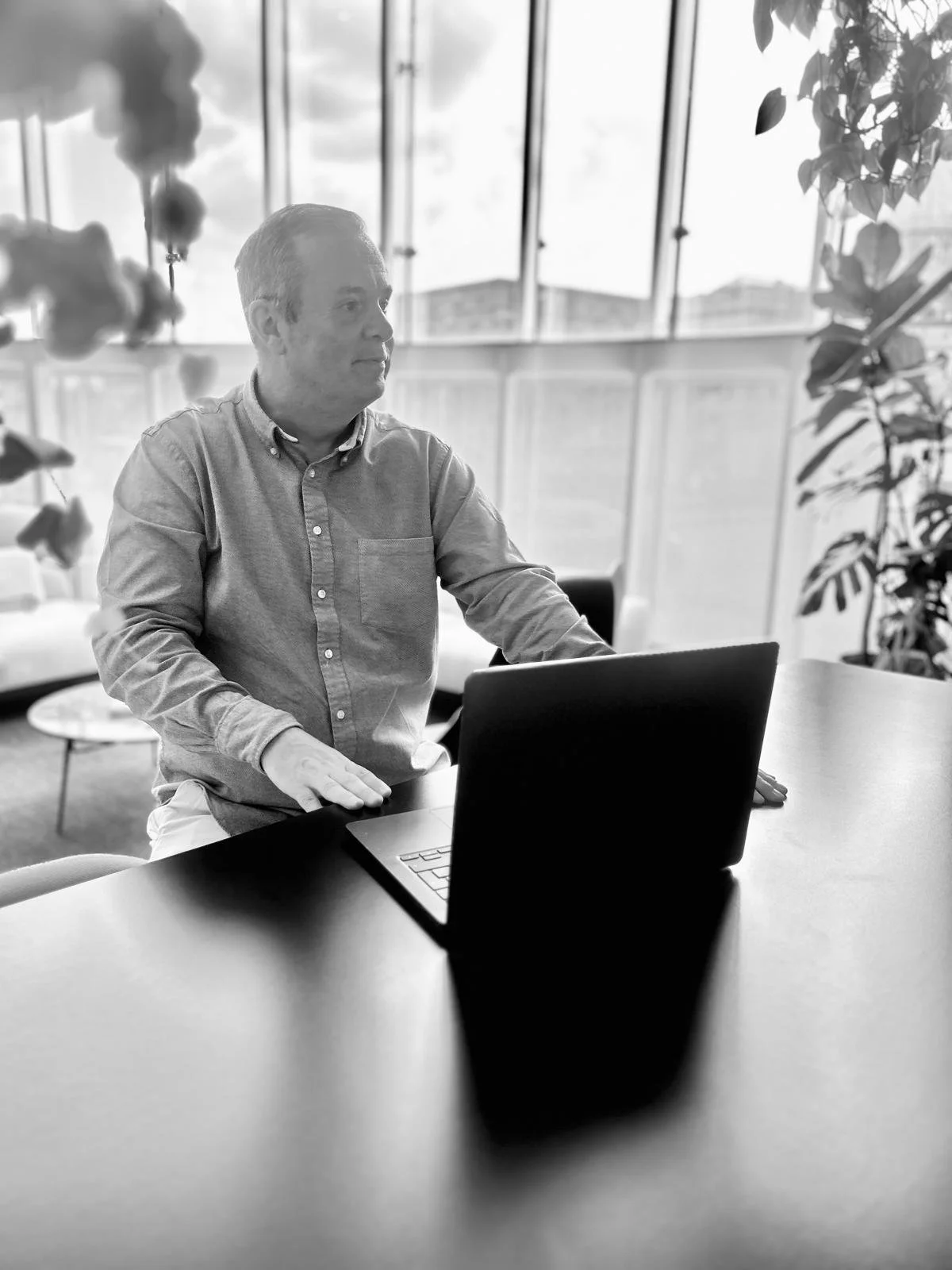 A man in a button-up shirt sitting at a table with a laptop, looking to his right, in a modern office setting with large windows and plants.