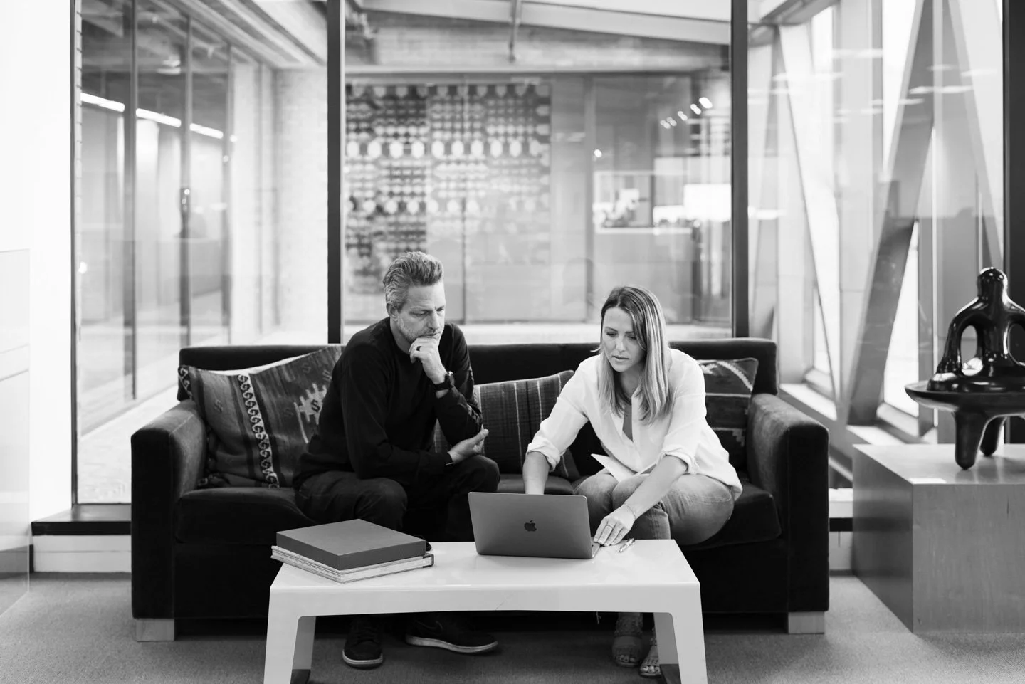 Two people sitting on a sofa looking at a laptop in what appears to be an office or lounge area, with a glass wall, books, and decorative objects around them.