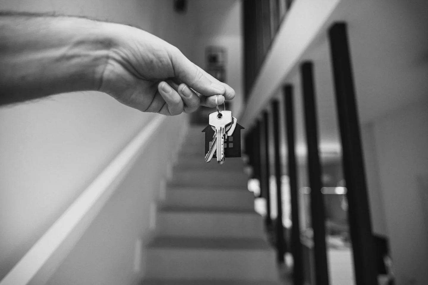 A hand holding up a keychain with house keys and a house-shaped key fob, in front of a staircase with black railings, in black and white.