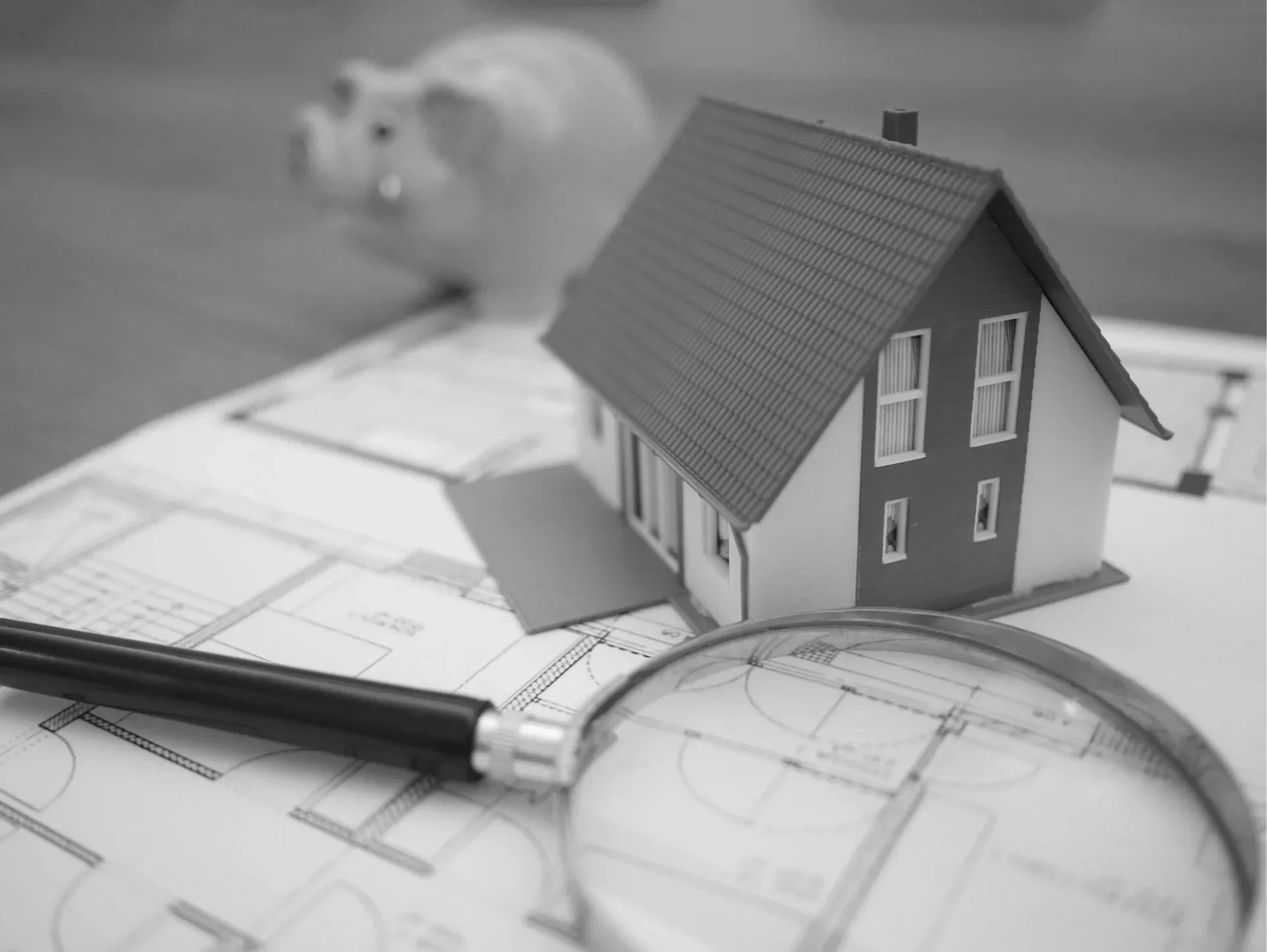 A miniature model house placed on architectural blueprints, with a black marker pen and a magnifying glass nearby, on a desk.