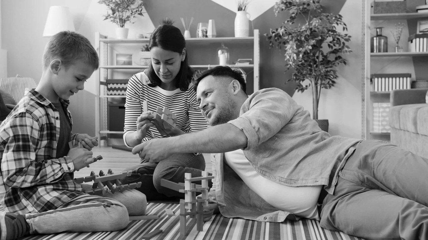 A family of three playing with wooden building blocks on a rug in their living room, smiling and enjoying each other's company.
