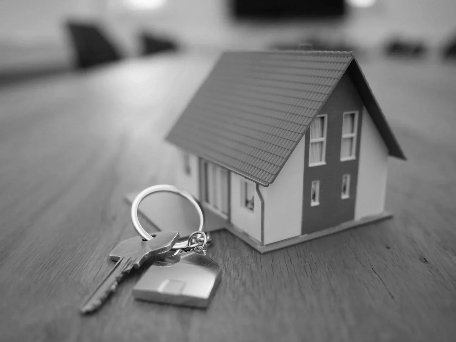 A small model house with a pitched roof and four windows, placed on a wooden surface. A keyring with keys is in the foreground, and the background shows a blurred interior setting.