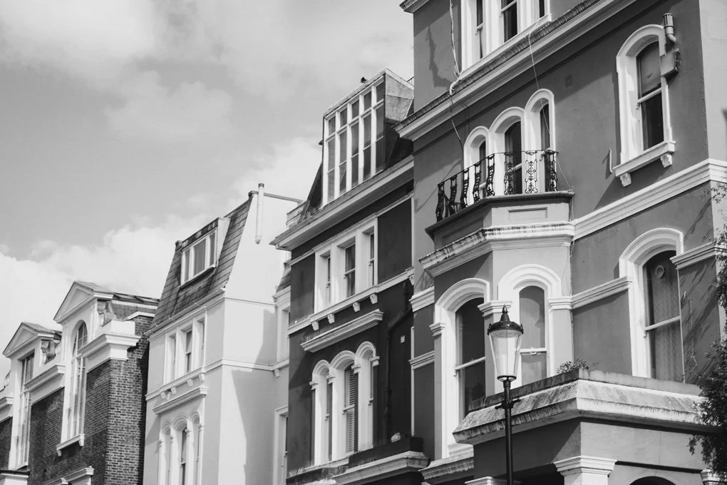 Row of multi-story residential buildings with ornate window frames, balconies, and a streetlamp in front, under a partly cloudy sky.